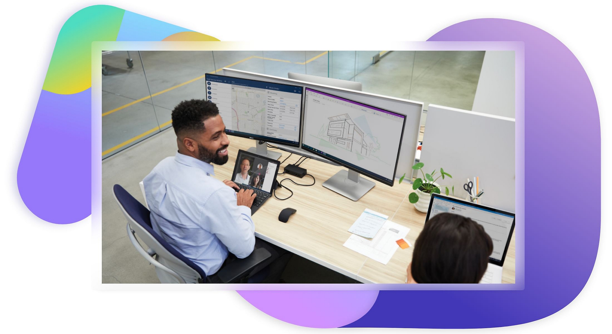 Một người đang làm việc trên máy tính xách tay và các màn hình máy tính tại bàn làm việc, đồng thời trò chuyện với đồng nghiệp ở bên cạnh.