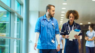 Two medical professionals walking down a hallway having a conversation and looking at a clipboard 