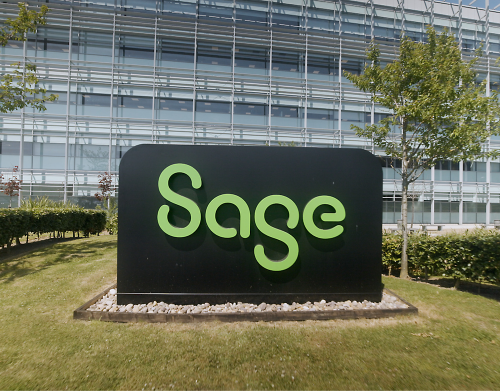 Un semn negru mare cu sigla verde Sage în fața unei clădiri moderne de birouri din sticlă, cu o amenajare peisagistică luxuriantă și verde.