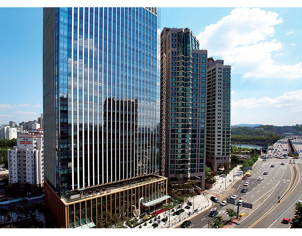 Modernes Stadtbild mit einem hohen gläsernen Bürogebäude neben Wohntürmen an einer belebten Straße unter einem klaren blauen Himmel.