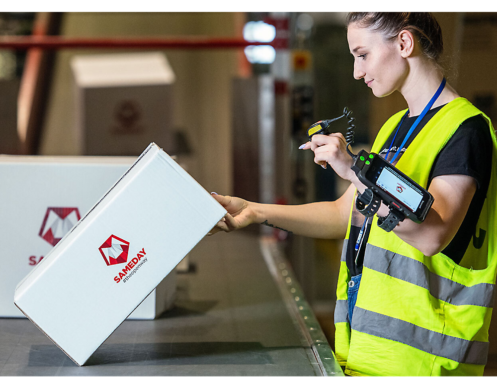 A female worker in a high visibility vest scans a barcode on a package in a warehouse.