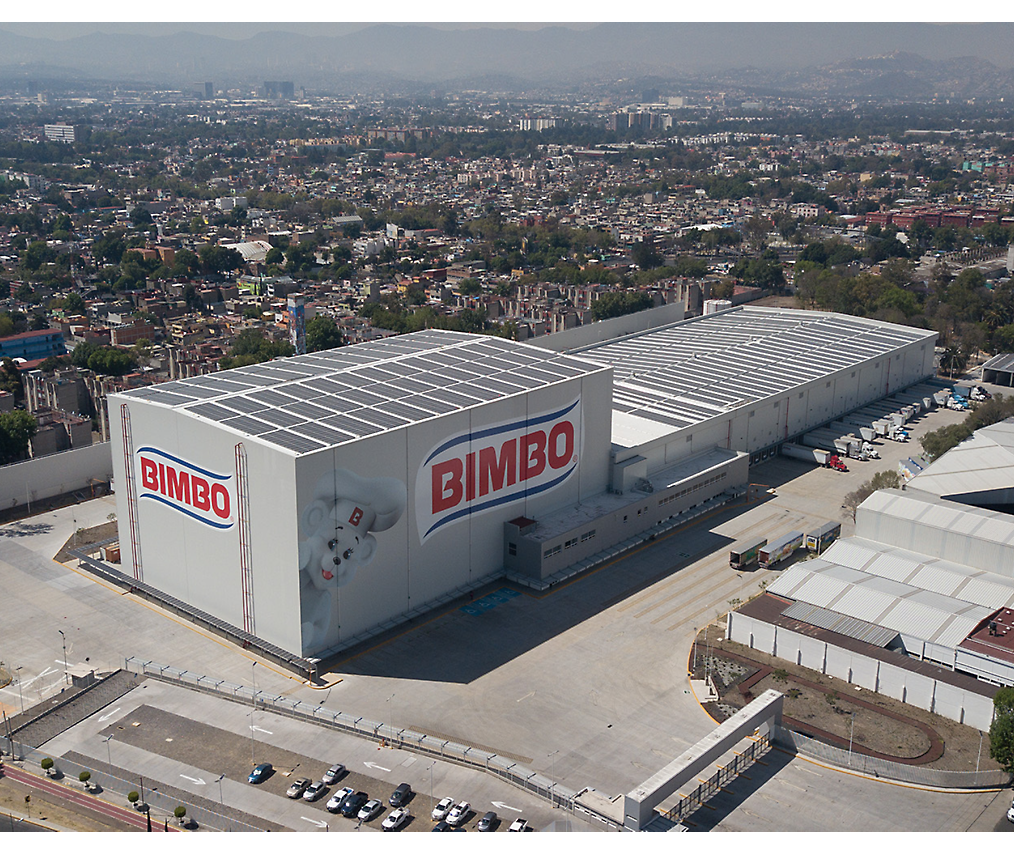 Aerial view of a large bimbo bakery facility with branded warehouses, surrounded by urban landscape.