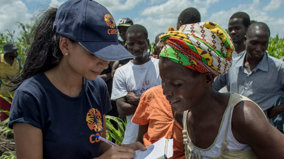 A woman writes notes while interacting with people in a field, surrounded by others.