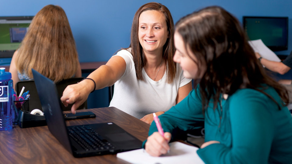 An instructor pointing to a laptop screen while a student looks on and takes notes.