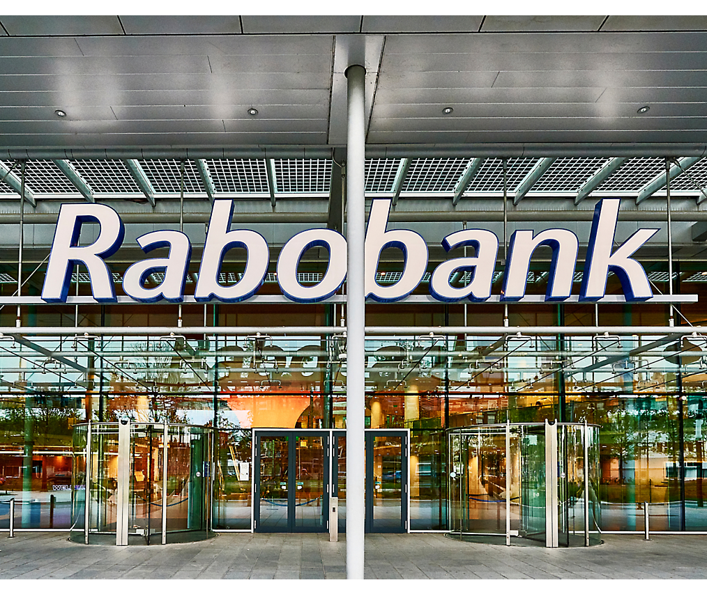 Buitenaanzicht van een Rabobank-gebouw met glazen deuren en een groot bord met de naam van de bank boven de ingang.