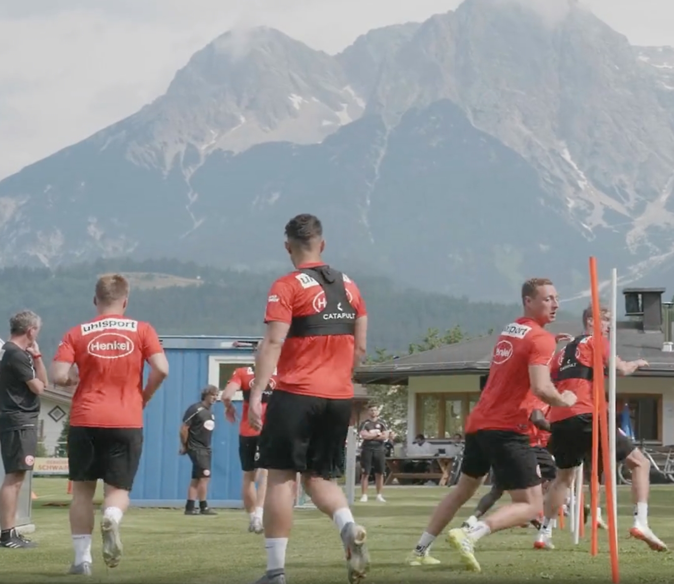 Fodboldspillere i røde trøjer træner udendørs med udsigt til bjerge i baggrunden.