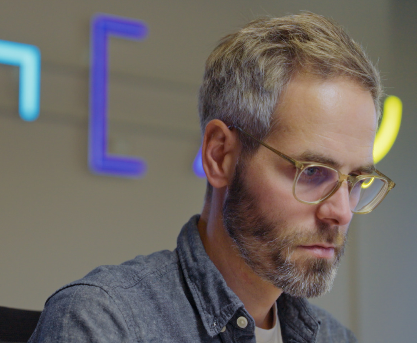 A man with a beard and glasses, focusing intently on a computer screen in an office with neon lights in the background.