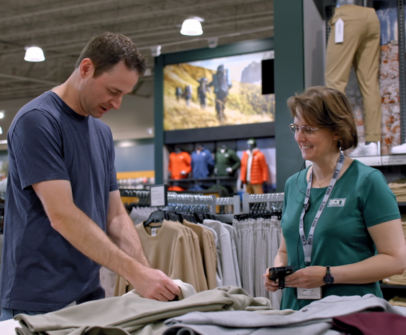 A man examines clothing at a store counter with assistance from a smiling female employee wearing a name tag.