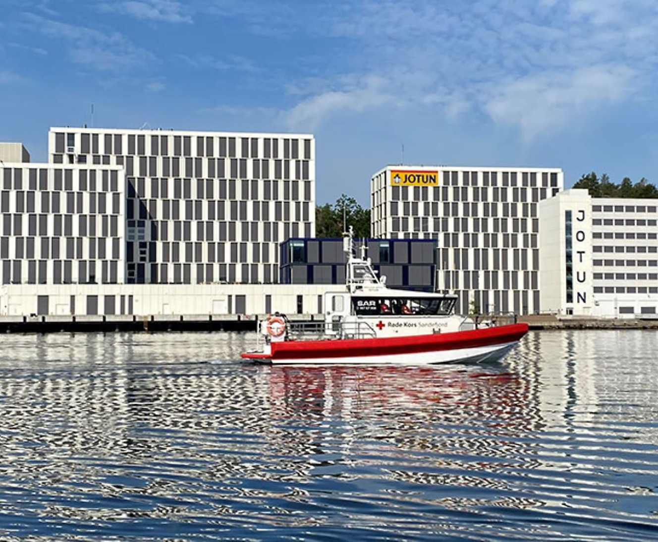 A red pilot boat on the water in front of modern waterfront buildings with "jotun" signage.