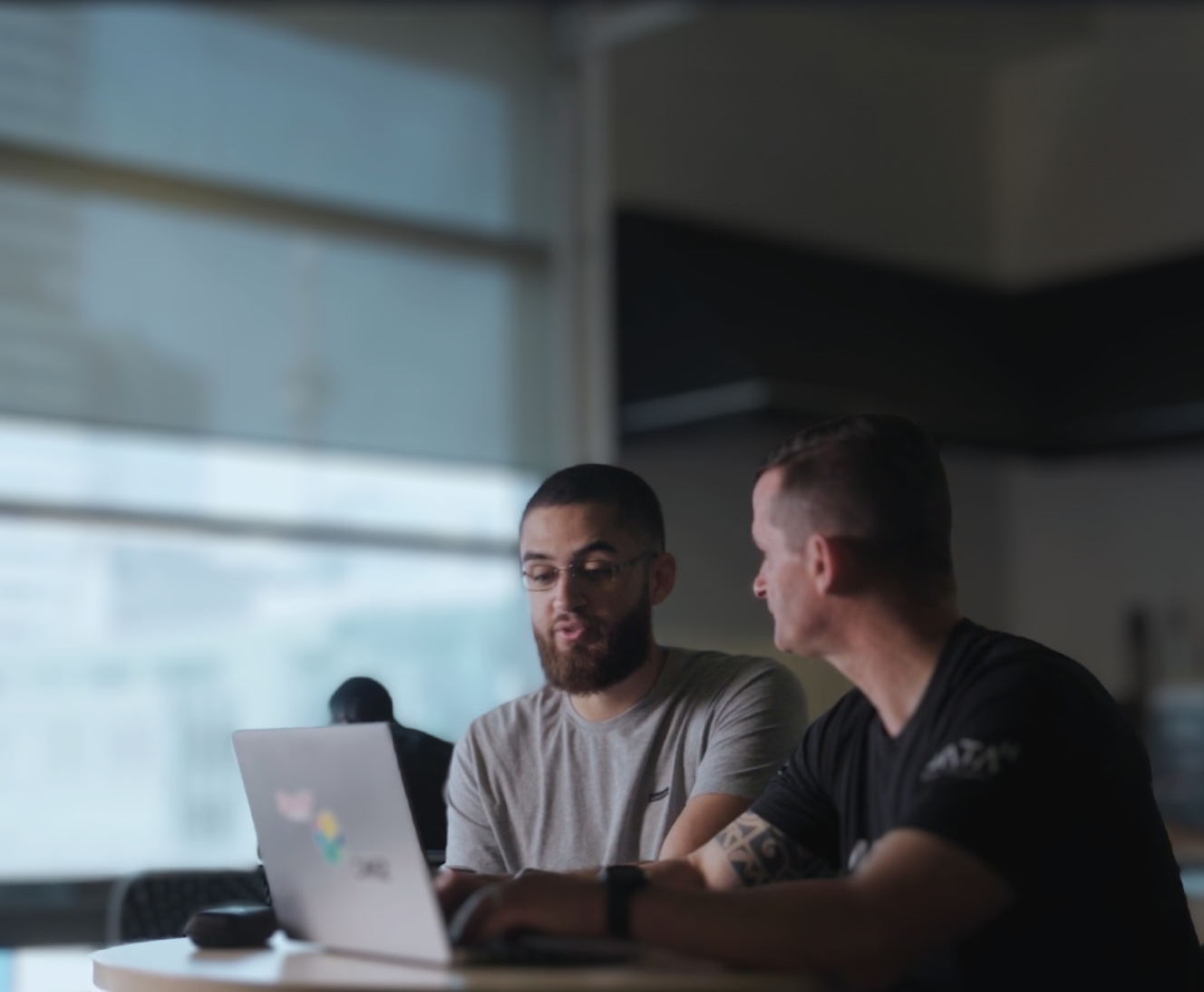 Two men, one with a beard, looking at a laptop screen together in a modern office setting with large windows.