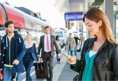 A woman checks her phone at a busy train station, with people walking by in the background.