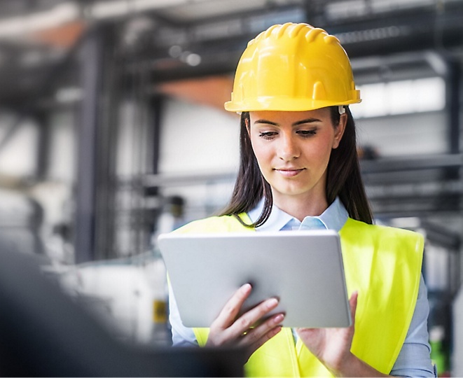 A woman wearing a hard hat is using a tablet in a factory.