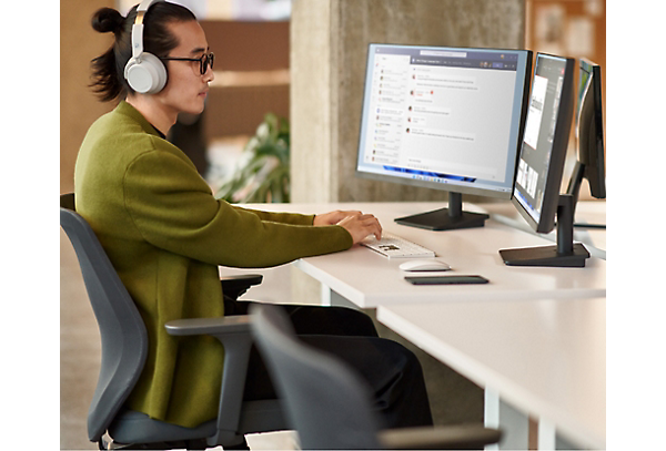 Osoba nosząca słuchawki i siedząca przy biurku z dwoma ekranami komputerowymi