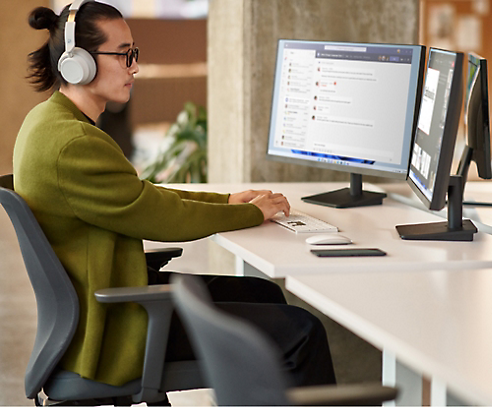 Een persoon die een hoofdtelefoon draagt en aan een bureau zit met twee computerschermen