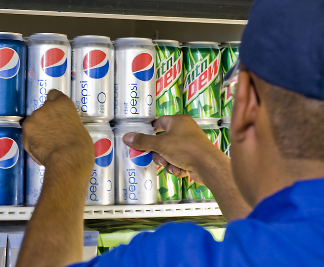 Um funcionário de loja reabastecendo latas de Pepsi e Mountain Dew em uma prateleira.