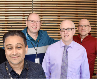 Four men posing for a photo in front of a wooden wall.
