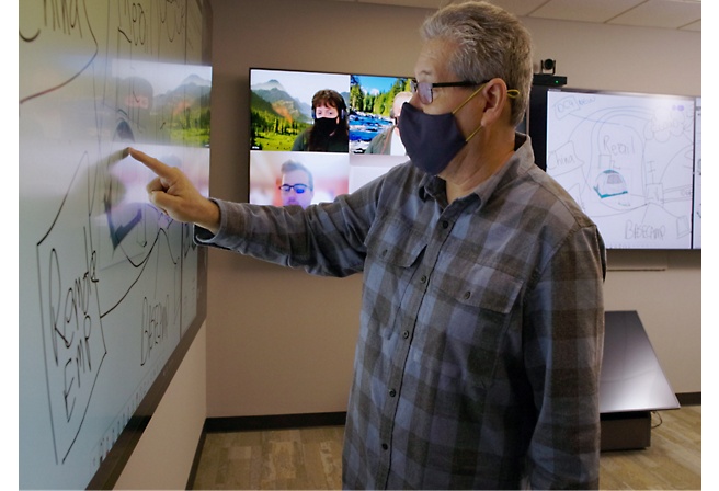 Man in a mask pointing at a whiteboard during a presentation.