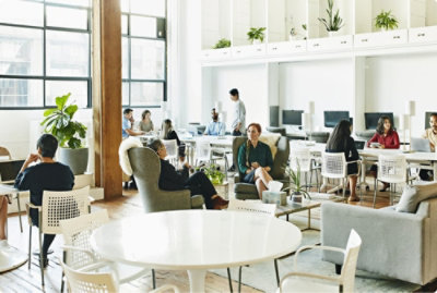 Modern office space with diverse employees working on laptops and discussing at tables, large windows, and ample greenery.