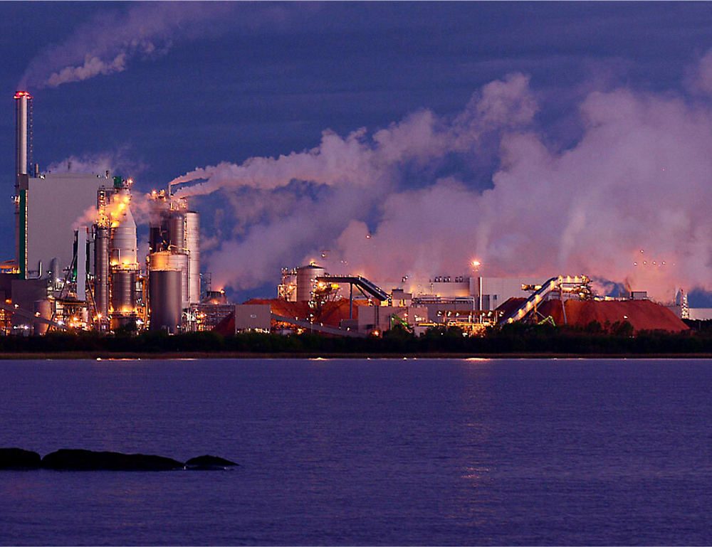 Industrieanlage mit mehreren Schornsteinen, die Rauch ausstoßen, in der Nähe eines Gewässers.
