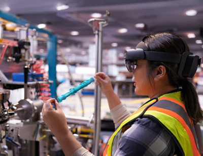 A person wearing augmented reality goggles and a safety vest examines a blue object in a manufacturing setting.