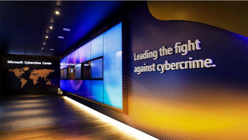Interior of the Microsoft Cybercrime Center featuring a world map labeled 'Microsoft Cybercrime Center' on the left, a large display screen with graphics and data in the center, and a wall on the right with the text 'Leading the fight against cybercrime.