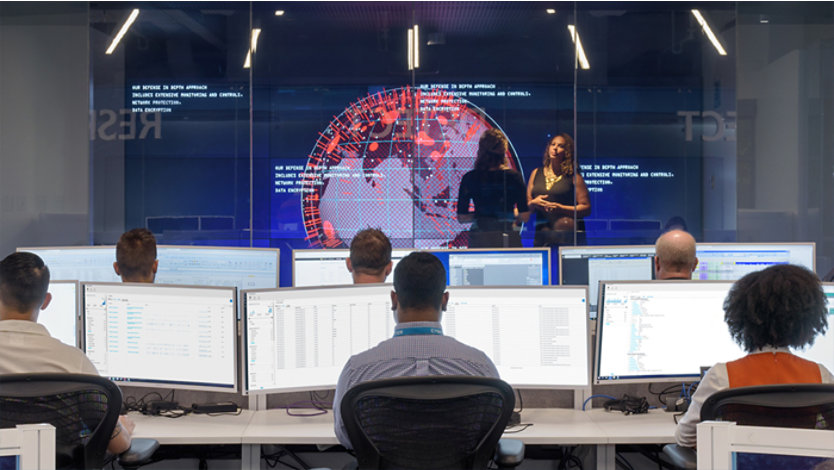 Office or control room with six people seated at computer stations, each with multiple monitors displaying data. In the background, a large screen shows a digital globe and the repeated text: 'Our defense in depth approach includes extensive detection and control measures to protect all environments.' Two individuals stand near the screen, appearing to discuss or present.