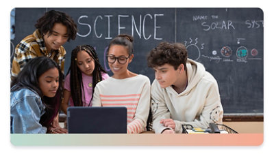 A teacher at her desk in a classroom surrounded by students, looking at a laptop screen.