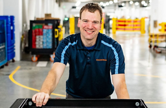 A Supported Employee, Sam, is smiling pushing a black cart through a warehouse. Stacks of boxes and materials are on the floor behind him.