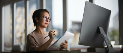 אישה מרכיבה משקפיים, מחזיקה עט ומחברת, מביטה בריכוז במסך מחשב בשטח משרדי מואר היטב.