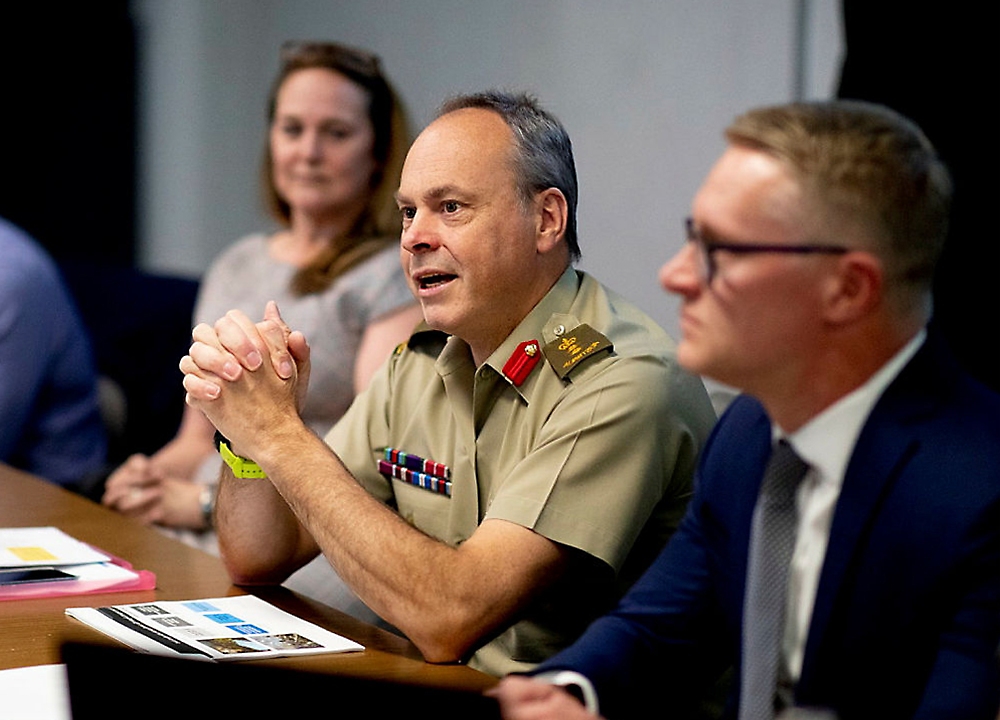 Un homme en uniforme militaire parle pendant une réunion avec deux autres participants attentifs dans une salle de conférence.