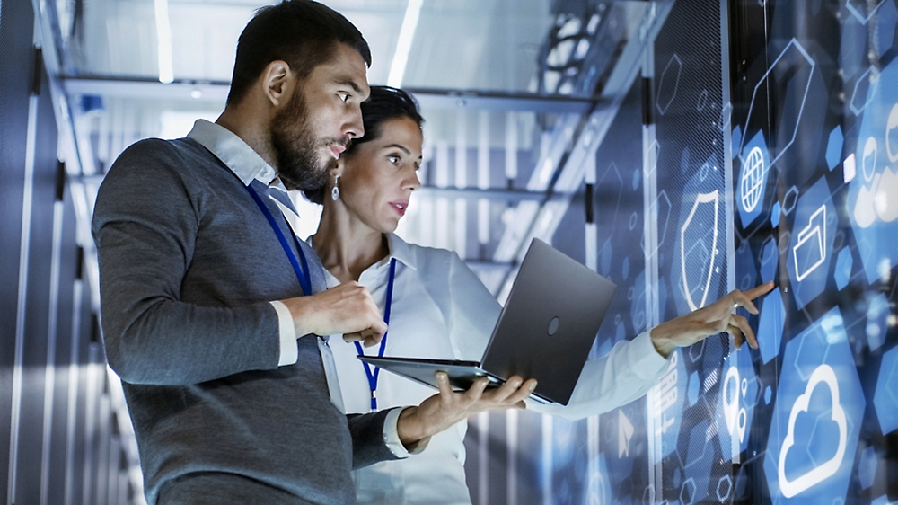 Dos profesionales analizando datos en pantallas en una sala de servidores de alta tecnología, uno sosteniendo un portátil