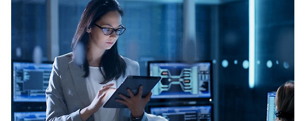 Seorang wanita mengenakan kacamata meninjau data pada tablet di ruang kontrol teknologi dengan cahaya redup.