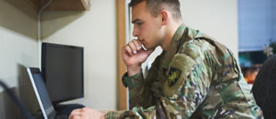 A person in military uniform focused on a laptop screen in an office setting.