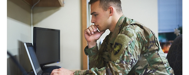 A person in military uniform focused on a laptop screen in an office setting.