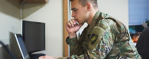 A person in military uniform focused on a laptop screen in an office setting.