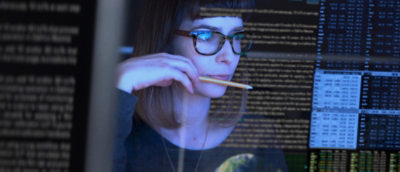 Woman with glasses examining code on multiple computer screens in a dark room, holding a pencil to her mouth.