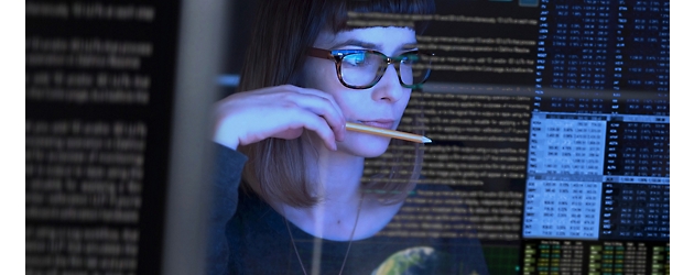 Woman with glasses examining code on multiple computer screens in a dark room, holding a pencil to her mouth.