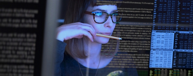 Woman with glasses examining code on multiple computer screens in a dark room, holding a pencil to her mouth.