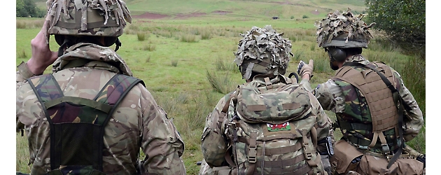 Drei Soldaten in Tarnkleidung stehen auf einem Feld, von der Kamera abgewandt, und beobachten die Landschaft.