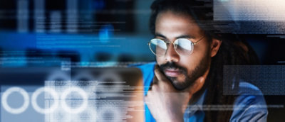 Man with long hair and glasses looking at computer screens displaying digital data overlays.