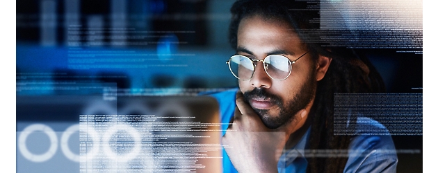 Un bărbat cu păr lung și ochelari privește ecrane de computer ce afișează suprapuneri de date digitale.