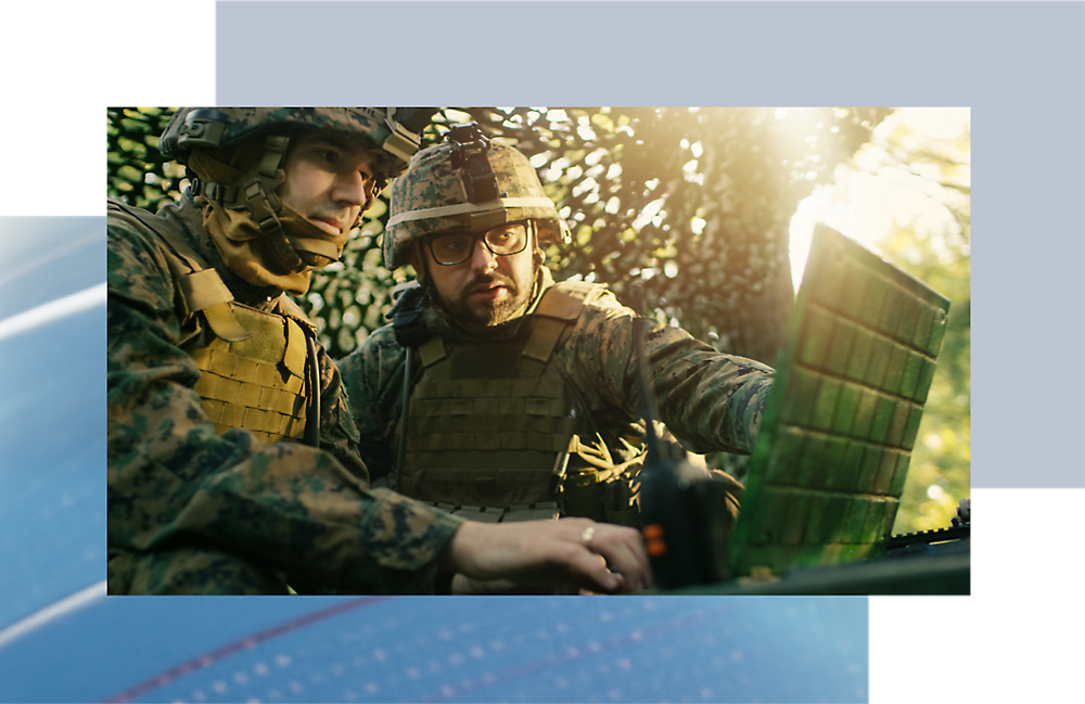 Deux soldats en camouflage en train d’examiner un ordinateur dans une forêt avec le soleil filtré par les feuilles.