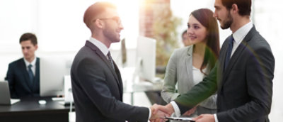 Two businesspeople shaking hands in a modern office environment, with another person working on laptop
