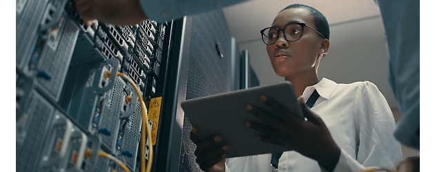 Uma mulher com óculos utiliza um tablet enquanto resolve problemas de hardware de servidor num datacenter.