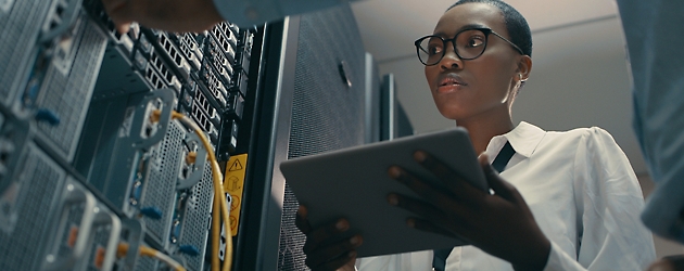 A woman with glasses uses a tablet while troubleshooting server hardware in a data center.