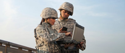 Two military personnel, a man and a woman, in camouflage uniforms using a portable device outdoors.