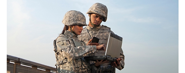 Two military personnel, a man and a woman, in camouflage uniforms using a portable device outdoors.