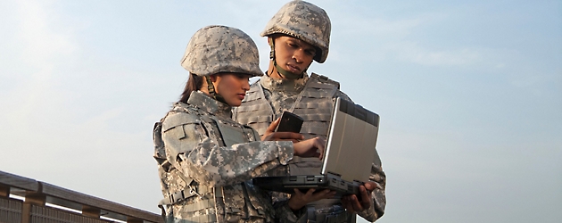 Two military personnel, a man and a woman, in camouflage uniforms using a portable device outdoors.