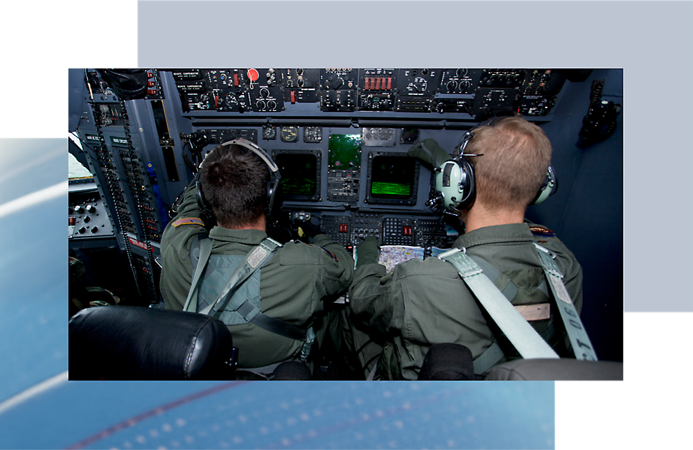 Two pilots operating controls in the cockpit of an aircraft, viewed from behind, with focus on the instrument panel.