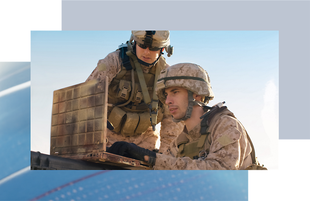 Two soldiers in combat gear examine a laptop outdoors under clear skies. one is standing and pointing at the screen.